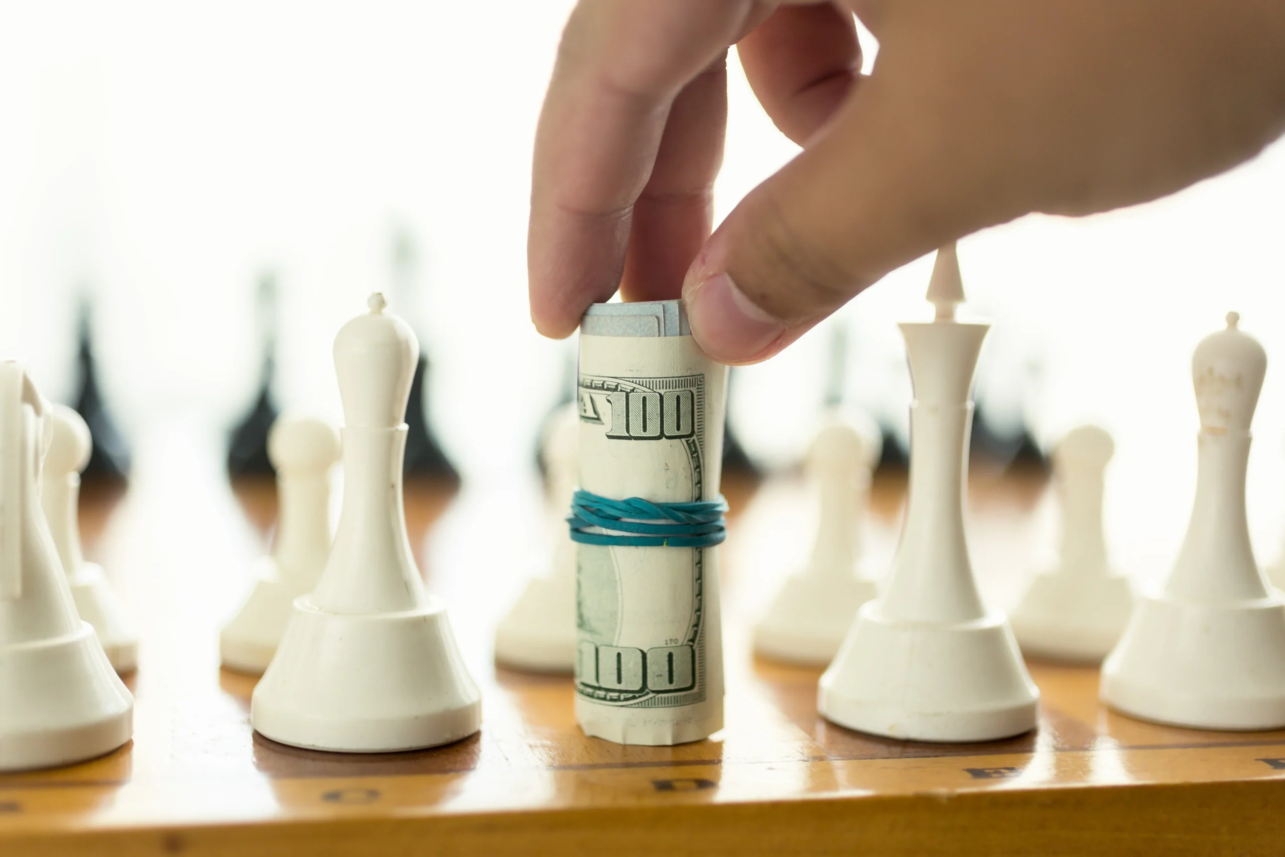 Hand moving a rolled-up U.S. $100 bill as a chess piece on a wooden chessboard, symbolizing financial strategy and investment decisions.
