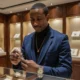 Mr. H in a jewelry showroom, inspecting a luxury wristwatch while wearing a navy blazer, black turtleneck, gold chain, and silver watch.